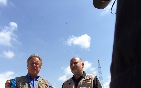 Two men wearing field/utility vests stand outdoors at what appears to be a disaster or construction site with debris-covered ground. The man on the left is speaking and holding a small blue object, while the man on the right wears a vest with a visible 'USAID' patch. Heavy machinery, cranes, and damaged or cleared structures are visible in the background.