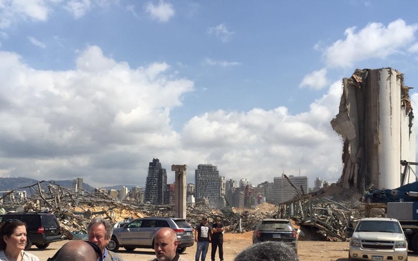 A group of officials and personnel are gathered at a heavily damaged port area, with massive destruction visible in the background including collapsed structures and a severely damaged grain silo tower. A crane branded 'HOUMANI' is visible on the right side. The scene appears to be a post-disaster assessment or briefing, consistent with the aftermath of the Beirut port explosion.