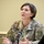 A U.S. Army female officer in OCP (Operational Camouflage Pattern) uniform is speaking at what appears to be a briefing or press conference. Her name tape reads 'DUGAN' and she wears a Colonel's eagle insignia along with a Medical Corps branch insignia patch on her sleeve. She is gesturing with both hands while speaking into a microphone.