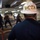 A Boatswain (BOSN) wearing a white hard hat with gold lettering oversees a crew of sailors working in what appears to be the interior deck of a naval vessel. The sailors in the background, wearing blue hard hats and navy uniforms, are engaged in a physical task involving ropes or lines and wooden materials. The scene suggests a damage control drill or deck seamanship training exercise aboard a military ship.