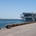 A large gray naval aircraft carrier is moored alongside a concrete pier or quay in what appears to be a military or commercial port. The vessel is viewed from the stern quarter, revealing its flight deck overhang, island superstructure, and various antennas and radar equipment. Industrial facilities and a long breakwater or pier structure are visible across the water in the background.