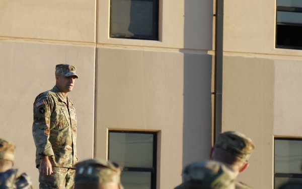 A military officer in US Army OCP (Operational Camouflage Pattern) uniform stands facing a formation of soldiers outside a tan/beige military building. The standing figure appears to be addressing or inspecting the assembled troops. The scene is consistent with a formal military formation, briefing, or ceremony.