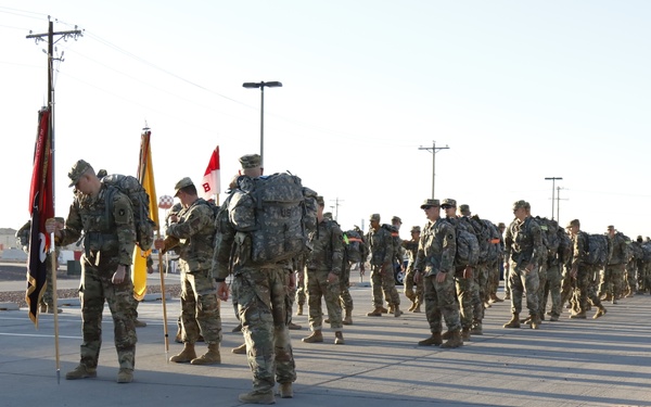 A large formation of military personnel in camouflage uniforms and full rucksacks are assembled on a wide paved road, likely on a military installation. Several soldiers near the front are carrying unit guidons and flags. The scene appears to be the beginning or staging of a ruck march or formation movement.