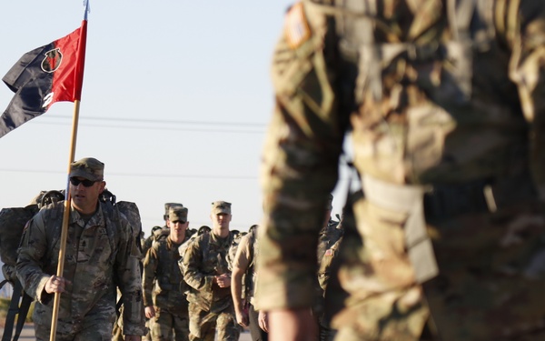 A column of U.S. Army soldiers in MultiCam/OCP camouflage uniforms conducts a road march or ruck march along a paved surface. A soldier near the front carries a unit guidon flag that is red and black with a circular emblem. The formation stretches back along the road with a military vehicle visible in the background.
