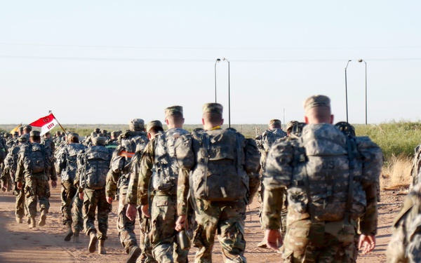 A large column of military personnel in camouflage uniforms and full rucksacks are conducting a road march along a dirt/gravel road in an arid, flat environment. The soldiers are moving away from the camera in a loose formation. A unit guidon flag is visible mid-column, suggesting this is an organized military ruck march or physical training event.