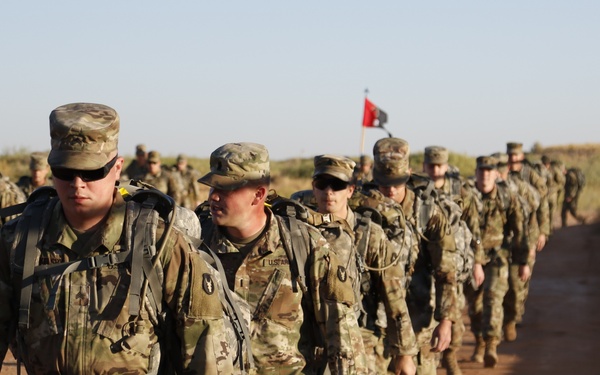 A column of U.S. Army soldiers in Operational Camouflage Pattern (OCP) uniforms conducts a road march along a dirt path in an arid, open terrain environment. The soldiers are carrying rucksacks and appear to be in formation, suggesting a training exercise or ruck march event. A red and black guidon flag is visible in the background, indicating unit identification.