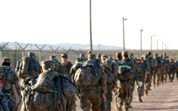 A large column of military personnel in camouflage uniforms conducting a ruck march along a dirt road in an arid, desert-like environment. The soldiers are carrying large rucksacks and wearing full combat gear, moving in a single-file or double-file formation. The setting appears to be a military installation, evidenced by the perimeter fencing with barbed wire visible on the left side.