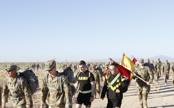 A large group of U.S. Army soldiers and personnel are conducting a ruck march along a dirt road in an arid desert environment. Some participants are in full combat uniform with rucksacks while others wear Army PT (physical training) black shorts and shirts. One individual is carrying what appears to be a unit guidon or ceremonial flag.