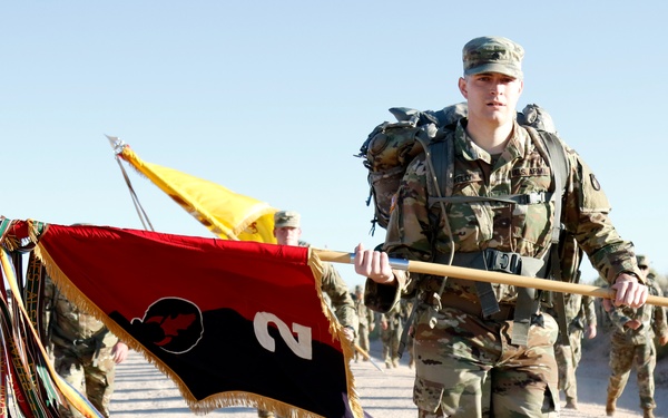 A U.S. Army soldier carries a unit guidon (flag) during what appears to be a ruck march or formation movement exercise. The primary flag is red and black with the number '2' and a buffalo/bison silhouette, adorned with multiple campaign streamers. A column of soldiers in full combat gear with rucksacks follows behind on a dirt road in an arid environment.