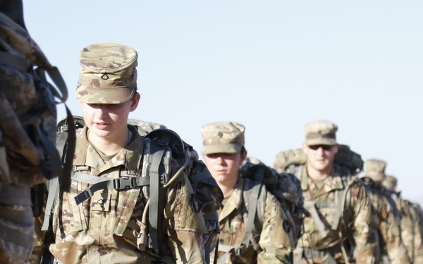 A column of U.S. Army soldiers in Operational Camouflage Pattern (OCP) uniforms conducts a ruck march across an arid, desert terrain. The soldiers are carrying large rucksacks and appear to be in formation, marching along a dirt road or trail. The image captures a training or field exercise environment, likely at a desert installation.