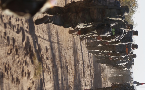 The image is rotated 90 degrees and shows a large column of individuals in military or paramilitary attire marching along a dirt road in an arid, dusty environment. At least one flag with red, white, and black coloring is visible among the group. The scene suggests a military patrol, convoy march, or organized movement operation.