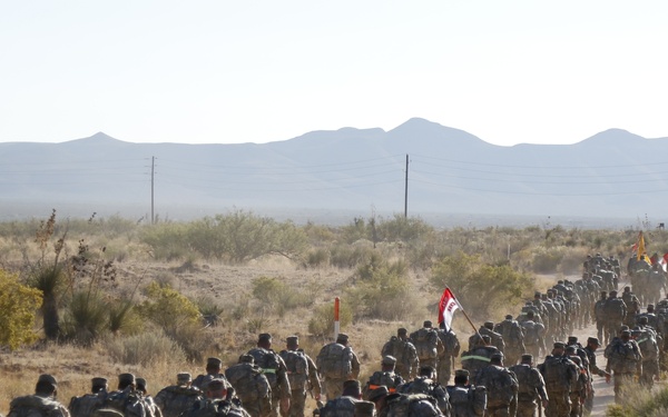 A large column of military personnel in camouflage uniforms and full rucksacks are conducting a road march along a dirt trail through an arid desert landscape. The formation stretches far into the distance, with at least one unit guidon flag visible mid-column. The terrain and vegetation suggest a southwestern United States desert environment, likely a military installation.