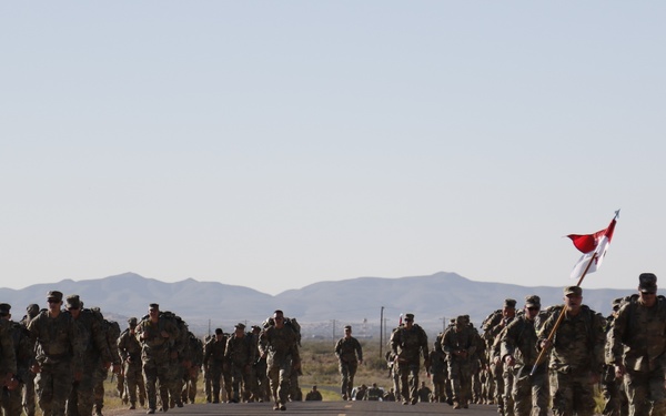 A large formation of military personnel in camouflage uniforms and full gear are conducting a road march along a two-lane desert highway. One soldier on the right side of the formation is carrying a red and white guidon flag. The terrain is arid and flat with mountains visible in the background under a clear sky.