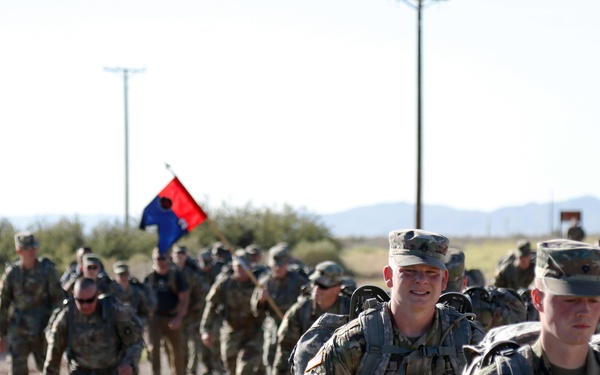A large column of U.S. Army soldiers in OCP (Operational Camouflage Pattern) uniforms conducts a road march along a paved road in an arid, desert-like environment. The soldiers are carrying rucksacks and a unit guidon flag (red and blue) is visible mid-column. The terrain and vegetation suggest a southwestern U.S. military installation, likely Fort Huachuca or a similar desert post.