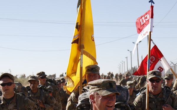 A large formation of U.S. Army soldiers in OCP (Operational Camouflage Pattern) uniforms conducting a ruck march along a dirt path beside a perimeter fence. Soldiers in the front carry unit guidons, including a yellow cavalry-style flag and red-and-white unit flags. The march appears to be taking place in an arid, desert environment consistent with a U.S. military installation.