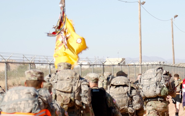 A column of U.S. Army soldiers in camouflage uniforms and full rucksacks marches along a dirt path beside a chain-link fence topped with razor wire. Two military unit guidons are visible — a yellow flag and a dark blue/red flag with what appears to be a unit insignia. The setting appears to be a military installation in an arid, desert environment.