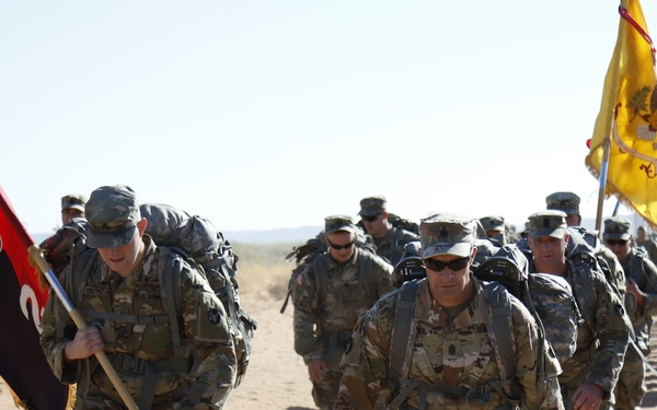 A column of military personnel in camouflage uniforms conducts a ruck march along a sandy desert road. Soldiers are carrying large backpacks and two guidon flags are visible — a red/black flag on the left and a yellow flag on the right. The setting appears to be an arid training environment, likely a U.S. Army installation.