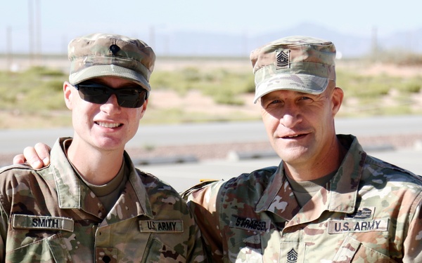 Two U.S. Army soldiers in Operational Camouflage Pattern (OCP) uniforms pose together outdoors at what appears to be a military installation in an arid/desert environment. The soldier on the left is identified by name tape as 'SMITH' and the soldier on the right appears to have a name tape reading 'STRASSER' or similar, wearing Sergeant Major rank insignia on his cap. Both are smiling for the photo in a friendly, informal pose.