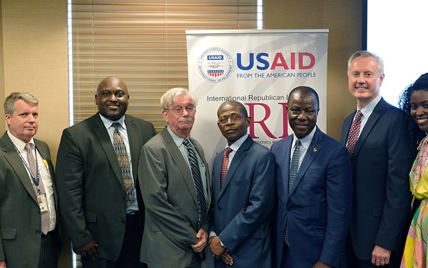 A group of approximately ten individuals pose for a formal group photograph in front of USAID and IRI (International Republican Institute) banners. A presentation screen in the background partially displays 'Sierra Leone Public C...' dated December 16, suggesting this is an official meeting or event related to Sierra Leone. The group includes both men and women dressed in a mix of Western business attire and traditional African clothing.