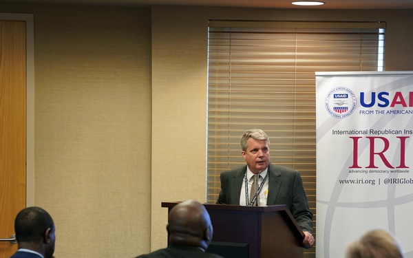 A man in a dark suit is speaking at a podium during a formal presentation or briefing. The event appears to be organized by the International Republican Institute (IRI) with funding from USAID, as indicated by the banner behind the speaker. An audience of approximately 6-8 attendees is seated facing the podium.