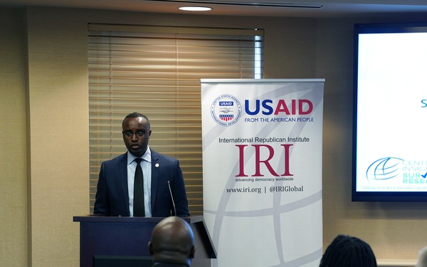A formal presentation or briefing is taking place in a conference room setting. A man in a dark suit is speaking at a podium, with banners from USAID and the International Republican Institute (IRI) visible behind him. The screen to the right displays a slide referencing 'Sierra Leone Public' and 'Center for Insights in Survey Research,' suggesting a policy or research briefing.