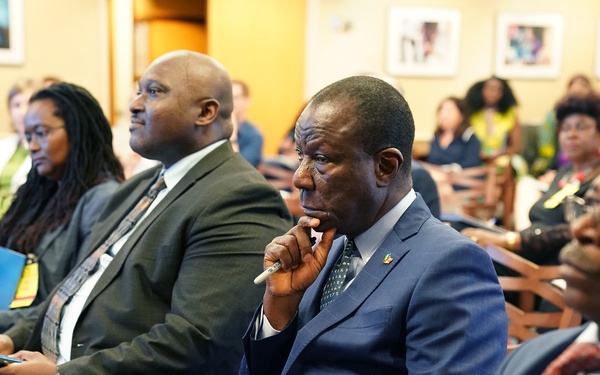 A formal meeting or conference is taking place in what appears to be a professional conference room or auditorium. Several attendees, predominantly dressed in business attire, are seated in rows listening attentively to a presentation or speaker. The central figure in a navy blue suit appears particularly engaged, holding a pen to his chin in a contemplative pose.
