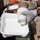 A person wearing white latex gloves and an orange high-visibility vest is handling disposable food containers, including a white foam clamshell takeout box and a paper cup labeled 'compostable'. The setting appears to be outdoors, possibly at a waste sorting or food distribution station. The image focuses on the hands and containers in the foreground.