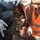 The image shows waste management workers handling garbage bags during a collection operation. At least two workers wearing high-visibility orange vests, white protective gloves, and face coverings are emptying a large black trash bag filled with dry leaves and organic debris into a collection vehicle. The scene appears to be an outdoor urban waste collection activity in bright daylight.