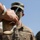 A close-up photograph showing a hand holding an inverted compostable cup with a straw, with military personnel in camouflage uniforms visible in the blurred background. The foreground subject wears a white wristband and a white sleeve. The setting appears to be outdoors under a clear blue sky.