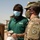 Two individuals are engaged in conversation in an outdoor military environment. One person is a U.S. Army soldier in OCP (Operational Camouflage Pattern) uniform with a 'SAPPER' tab and an 'A' unit patch, wearing a camo face mask and cap with a partially visible name tag reading 'KESS--'. The other individual is a civilian wearing a green Vectrus company polo shirt and a blue surgical mask, holding a paper cup and bag.
