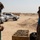 A civilian man in a green polo shirt speaks with a U.S. Army soldier in MultiCam uniform in an arid, desert environment. Both individuals are wearing face masks, suggesting this was taken during the COVID-19 pandemic period. The setting appears to be a forward operating base or military installation, with sandbag barriers and white vehicles visible in the background.