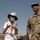 Two individuals are standing outdoors in what appears to be a military installation or forward operating base in an arid environment. One person is a U.S. Army captain in OCP (Operational Camouflage Pattern) uniform, and the other is a civilian wearing a white shirt, bucket hat, and lanyard with credentials. Both are wearing face masks, suggesting this was taken during the COVID-19 pandemic period.
