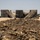 The image shows a close-up foreground of a mound of dry, dark soil mixed with organic debris such as twigs and straw. In the blurred background, there are large cylindrical structures that appear to be collapsed or damaged storage tanks or silos, set in an arid, open landscape. The scene suggests a rural or industrial site in a dry climate.
