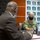 Two individuals are seated at a conference table in what appears to be an official meeting setting. One person is wearing U.S. Navy camouflage uniform with a black face mask and a name tape reading 'SCHULMAN', while the other individual, seen from behind, is wearing a brown suit with a blue face mask. The backdrop displays repeated logos and text reading 'Ministère du Travail' (Ministry of Labor), suggesting a formal bilateral or diplomatic meeting.