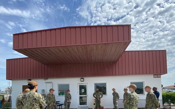 A group of military personnel in U.S. Navy Working Uniform (NWU Type III, woodland camouflage pattern) are gathered outside a low-profile building numbered '3070' on what appears to be a military installation. The personnel appear to be assembled in a loose formation or briefing configuration in an outdoor courtyard or common area. The setting suggests a routine muster, briefing, or inspection activity at a naval base facility.