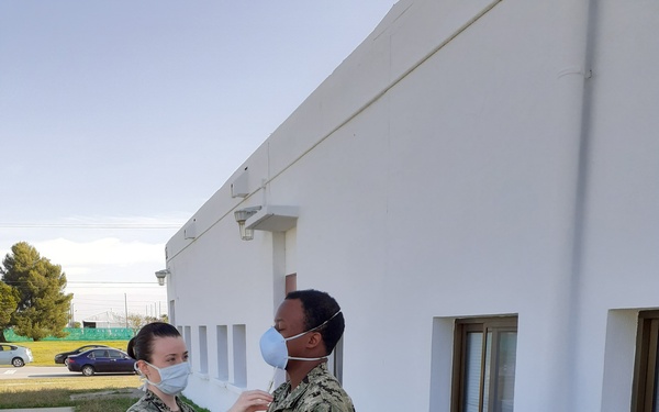 Two U.S. Navy personnel in NWU Type III (digital camouflage) uniforms are standing outdoors near a white building. One service member appears to be pinning or adjusting something on the collar/chest area of the other, suggesting a promotion or award ceremony. Both individuals are wearing light blue surgical face masks, consistent with COVID-19 era protocols.