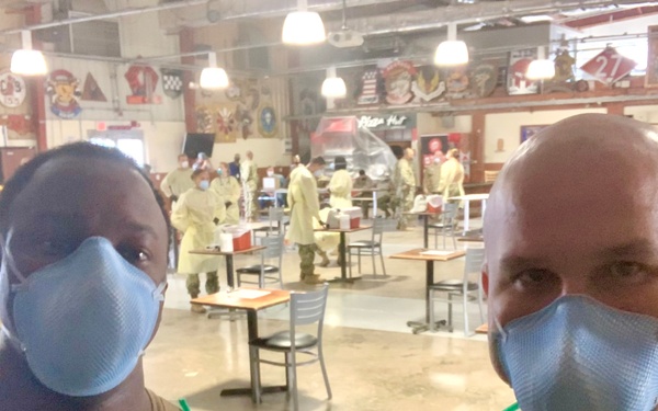 Two individuals wearing yellow disposable gowns, blue face masks, and gloves pose for a selfie inside what appears to be a military dining facility (DFAC) converted into a medical screening or vaccination site. In the background, multiple personnel in similar PPE are stationed at tables, suggesting an organized medical operation. A Pizza Hut sign and various military unit insignia/patches are visible on the walls.