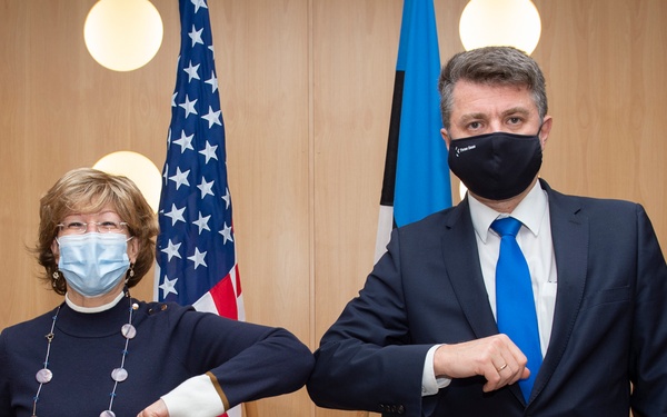 Two individuals wearing face masks are performing an elbow bump greeting, a COVID-19 era alternative to handshakes. They are standing in front of an American flag and what appears to be an Estonian flag, suggesting an official diplomatic or governmental meeting. The setting includes framed photographs on a wood-paneled wall, indicating a formal office environment.