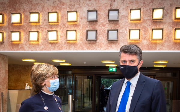 Four individuals are gathered in what appears to be a formal institutional building lobby, all wearing face masks consistent with COVID-19 pandemic protocols. They appear to be engaged in a conversation or meeting, dressed in professional business attire. The setting features a distinctive wall with illuminated square light fixtures in a grid pattern.