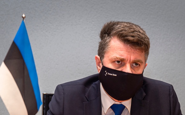 A man in a dark suit and blue tie sits at a desk wearing a black face mask labeled 'Three Seas'. An Estonian flag (blue, black, and white horizontal stripes) is visible on the desk to his left. The setting appears to be a formal or official meeting environment.