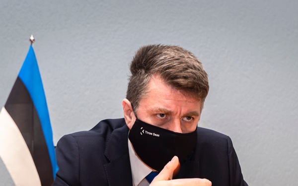 A man in a dark suit and blue tie is seated at a table, wearing a black face mask with the text 'Three Seas' printed on it. He is gesturing with his hands in what appears to be a formal or diplomatic setting. A small Estonian flag (blue, black, and white horizontal stripes) is visible on the table beside him.