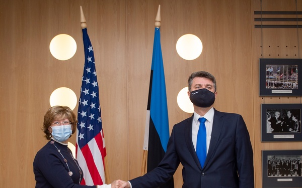 Two individuals wearing face masks are shaking hands in a formal diplomatic setting, standing in front of the United States flag and the Estonian flag. The setting appears to be an official government or diplomatic venue, decorated with framed photographs on a wood-paneled wall. The meeting likely represents a bilateral diplomatic engagement between the United States and Estonia.