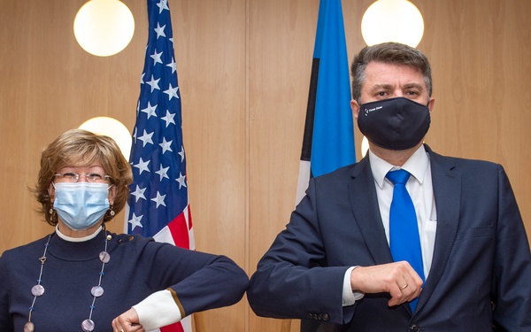 Two masked individuals are performing an elbow bump greeting, a COVID-19 era alternative to handshakes, in what appears to be an official government or diplomatic setting. An American flag and what appears to be an Estonian flag are visible in the background, suggesting a bilateral diplomatic meeting. The setting includes framed photographs on wood-paneled walls, indicating a formal official environment.
