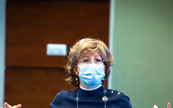 A woman wearing a blue surgical mask and dark navy top with a beaded necklace is seated at a conference table, gesturing with both hands as if speaking or presenting. The setting appears to be a formal meeting or discussion, likely during the COVID-19 pandemic period. Two other individuals are partially visible in the foreground, seated across from her.