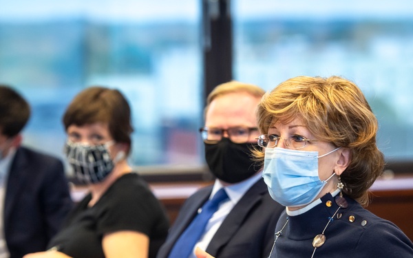 A formal meeting or conference setting where four individuals are seated at a dark conference table, all wearing face masks consistent with COVID-19 era protocols. The woman in the foreground, wearing a navy outfit with a decorative necklace, appears to be actively speaking and gesturing with her hands. The background shows large windows with an urban view, suggesting a high-rise office or conference facility.