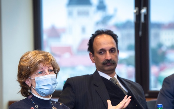 Three individuals are seated at a conference table in what appears to be a formal meeting setting. A woman in the foreground is speaking and gesturing with both hands while wearing a blue surgical mask. Two men are seated behind her, one without a mask and one wearing a white surgical mask.