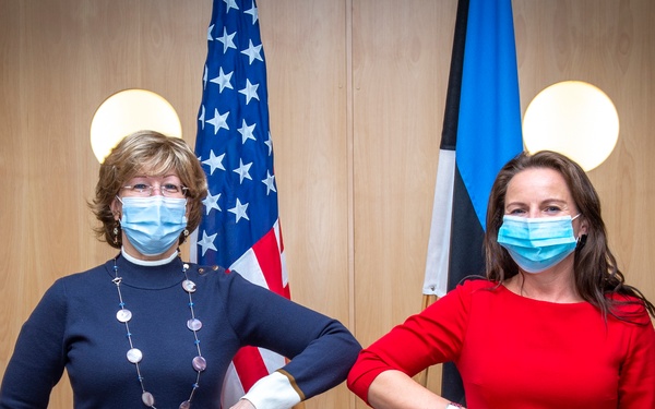 Two women wearing blue surgical face masks are posing together in what appears to be an official or diplomatic setting, performing an elbow bump greeting instead of a handshake. They are standing in front of an American flag and what appears to be an Estonian flag, suggesting a diplomatic or official meeting context. The setting includes framed photographs on a wood-paneled wall.