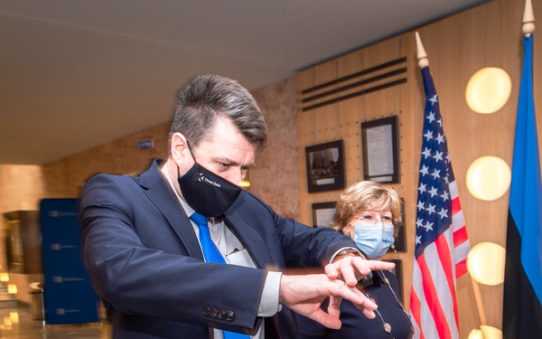 The image shows three individuals in a formal indoor setting, all wearing face masks, suggesting a COVID-era official or diplomatic meeting. Two flags are prominently displayed in the background — the United States flag and the Estonian flag — indicating a bilateral or diplomatic context. A man in a navy suit with a blue tie appears to be gesturing or directing attention toward something, while a woman in a dark jacket stands nearby.
