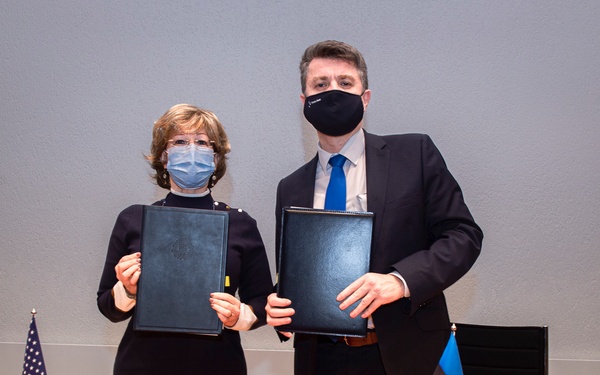 Two individuals wearing face masks are posing at a conference table, each holding a dark leather document folder, suggesting the signing of an official agreement or treaty. A small American flag and what appears to be the Estonian flag (blue, black, and white horizontal stripes) are visible on the table, indicating a bilateral diplomatic event. A colorful floral arrangement serves as a centerpiece on the wooden conference table.
