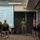 A military briefing or training session is taking place in a conference room, with one soldier standing at the front presenting to a small group of seated personnel. A projected presentation slide titled 'Priorities' is visible on the screen behind the presenter. All individuals are wearing camouflage uniforms and face masks, suggesting this was taken during the COVID-19 pandemic period.